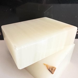 May include: Two rectangular, off-white blocks of stone, one resting atop the other. The top block is smooth and solid, while the bottom block has a small, textured, brown inclusion. The blocks are angled, creating a dynamic composition.