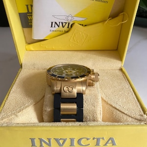 May include: A gold-toned Invicta watch with a black band rests in a yellow box. The watch has a green face and a gold bezel. The box contains an instruction manual and warranty information.