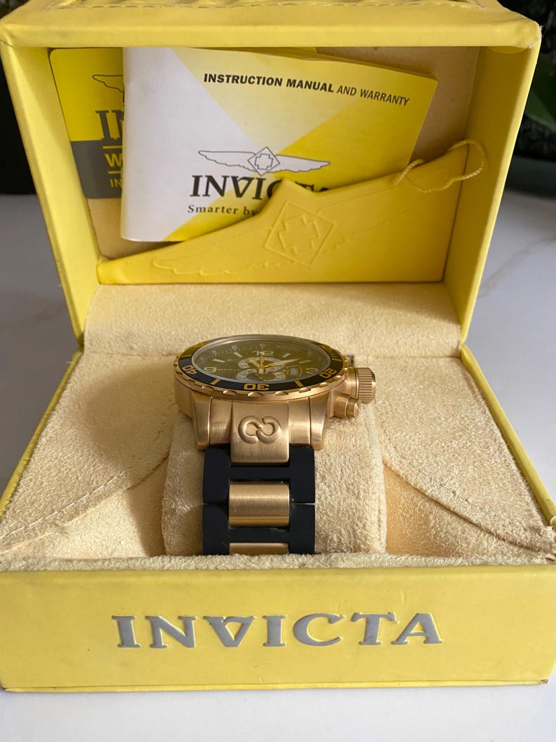 May include: A gold-toned Invicta watch with a black band rests in a yellow box. The watch has a green face and a gold bezel. The box contains an instruction manual and warranty information.