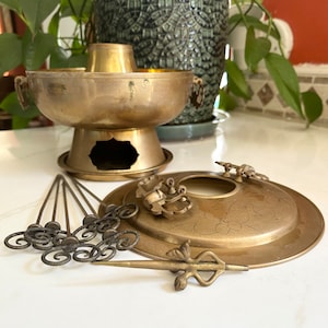 May include: A brass hot pot set with a round pot, a base, a lid, and six skewers. The pot has a floral design on the lid and a handle on each side. The base has a hole in the center. The skewers are thin and have a spiral design at the end.