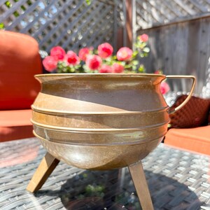 Op de afbeelding: Een bronzen metalen ketel met drie poten en een handvat. De ketel heeft horizontale banden rondom de body. De achtergrond omvat een houten hek, rode kussens en roze bloemen.
