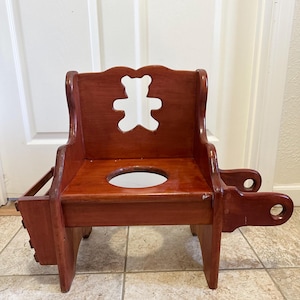 May include: A reddish-brown wooden potty chair. It has a bear-shaped cutout in the backrest and a round opening in the seat. The chair also features a small storage compartment and a classic design. The chair is made of wood.