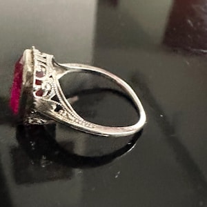 Op de afbeelding: Een zilveren ring met een rechthoekige, dieproze edelsteen. De ring heeft ingewikkelde filigraan details rond de edelsteen en langs de band, wat wijst op een vintage of antieke stijl. De ring staat tegen een donkere achtergrond.