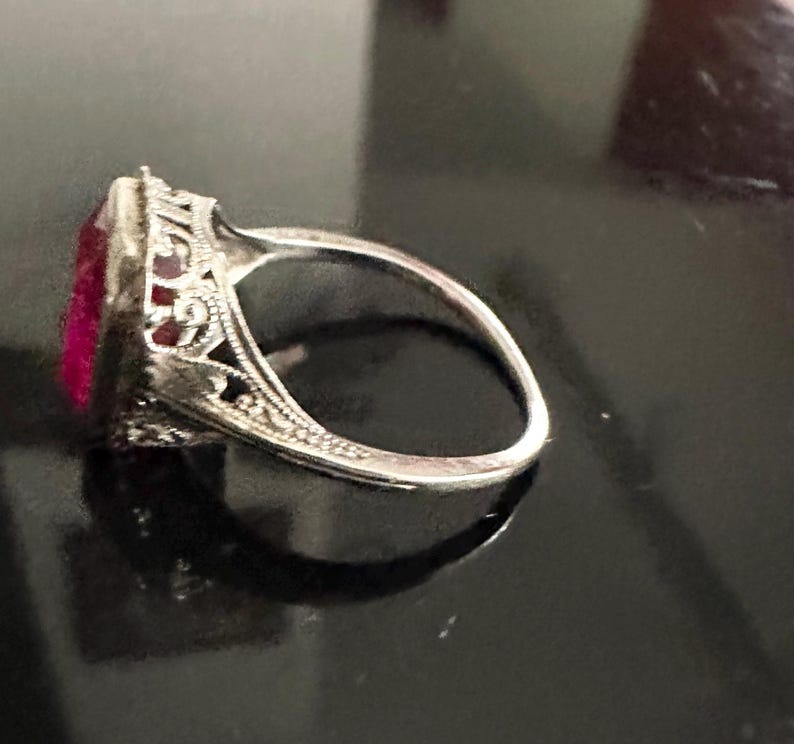 Op de afbeelding: Een zilveren ring met een rechthoekige, dieproze edelsteen. De ring heeft ingewikkelde filigraan details rond de edelsteen en langs de band, wat wijst op een vintage of antieke stijl. De ring staat tegen een donkere achtergrond.