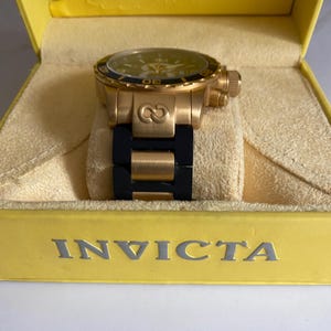 May include: A gold-toned Invicta watch with a black band, displayed in a yellow box. The watch features a black bezel and a yellow face. The watch is a luxury item.