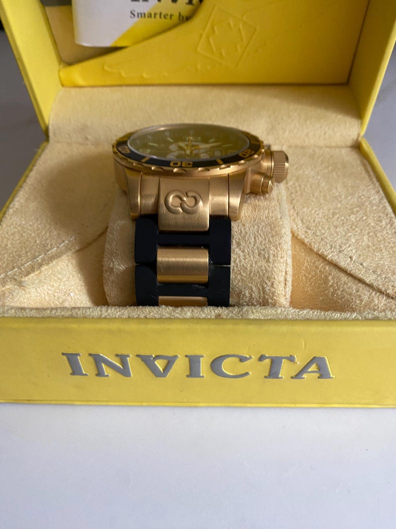 May include: A gold-toned Invicta watch with a black band, displayed in a yellow box. The watch features a black bezel and a yellow face. The watch is a luxury item.