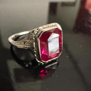 Op de afbeelding: Een zilveren ring met een rechthoekige, dieprode edelsteen. De ring heeft een sierlijk filigraanontwerp rond de edelsteen en op de band. De edelsteen reflecteert licht, waardoor een sprankelend effect ontstaat. Sieraden.