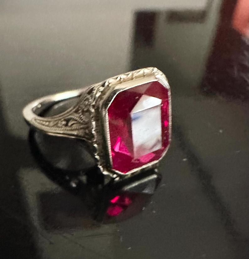 Op de afbeelding: Een zilveren ring met een rechthoekige, dieprode edelsteen. De ring heeft een sierlijk filigraanontwerp rond de edelsteen en op de band. De edelsteen reflecteert licht, waardoor een sprankelend effect ontstaat. Sieraden.