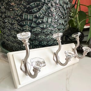 Puede incluir: Un perchero de pared blanco con cuatro ganchos decorativos. Cada gancho presenta un pomo de cristal transparente y facetado y una base de metal de níquel cepillado curvada. El perchero es rectangular y está frente a una maceta de cerámica verde oscuro.