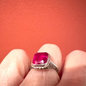 Op de afbeelding: Een vintage zilveren ring met een grote, vierkant geslepen, helderrode edelsteen. De ring heeft een sierlijk filigraanontwerp op de band en rond de edelsteen setting. De achtergrond is een effen, warme rode kleur.
