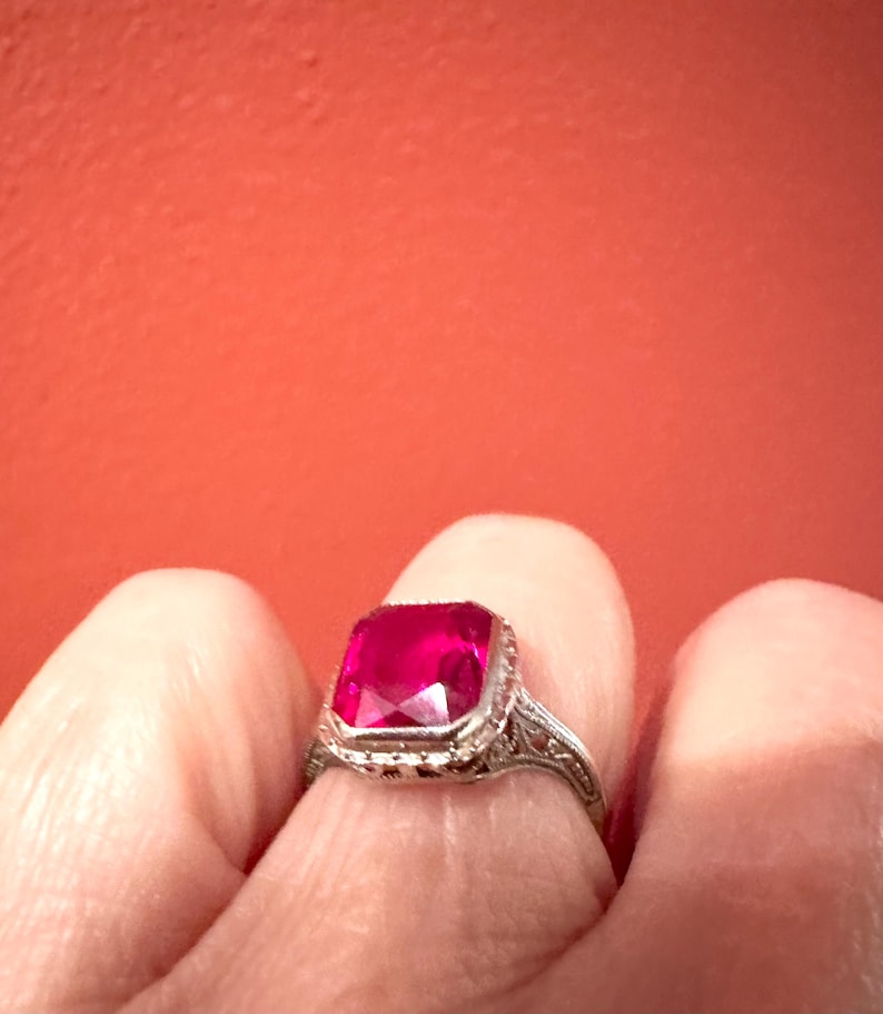 Op de afbeelding: Een vintage zilveren ring met een grote, vierkant geslepen, helderrode edelsteen. De ring heeft een sierlijk filigraanontwerp op de band en rond de edelsteen setting. De achtergrond is een effen, warme rode kleur.
