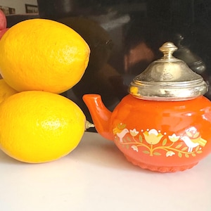 Op de afbeelding: Drie heldergele citroenen naast een oranje theepot met een zilveren deksel. De theepot is versierd met een bloemen- en vogelontwerp in wit en geel. De theepot heeft een gebogen tuit en handvat.