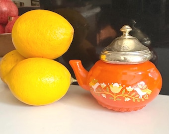 Puede incluir: Tres limones amarillos brillantes junto a una tetera naranja con tapa plateada. La tetera está decorada con un diseño floral y de pájaros en blanco y amarillo. La tetera tiene un pico y un asa curvados.