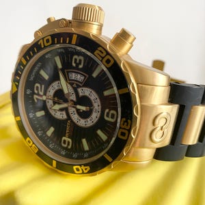 May include: Gold-toned Invicta chronograph watch with a black dial and bezel. Features include a tachymeter, date display, and luminous hands. The watch has a gold band and a black rubber strap.