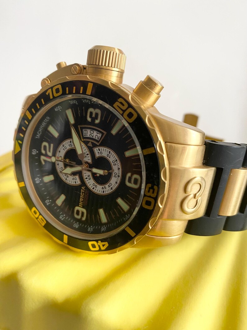 May include: Gold-toned Invicta chronograph watch with a black dial and bezel. Features include a tachymeter, date display, and luminous hands. The watch has a gold band and a black rubber strap.