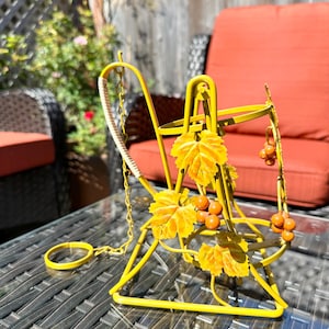 Puede incluir: Un soporte para botellas de vino de metal amarillo con detalles de uvas y hojas. El soporte tiene una cadena y un anillo para colgar. El diseño incluye hojas amarillas y racimos de uvas naranjas. El soporte está sobre una mesa de cristal.