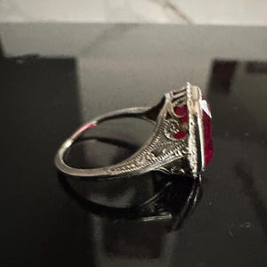 Op de afbeelding: Een zilveren ring met een sierlijk filigraanontwerp en een grote, rechthoekige, dieprode edelsteen. De ring is geplaatst tegen een donkere achtergrond, waardoor de ingewikkelde details van het metaalwerk en de levendige kleur van de steen worden benadrukt.