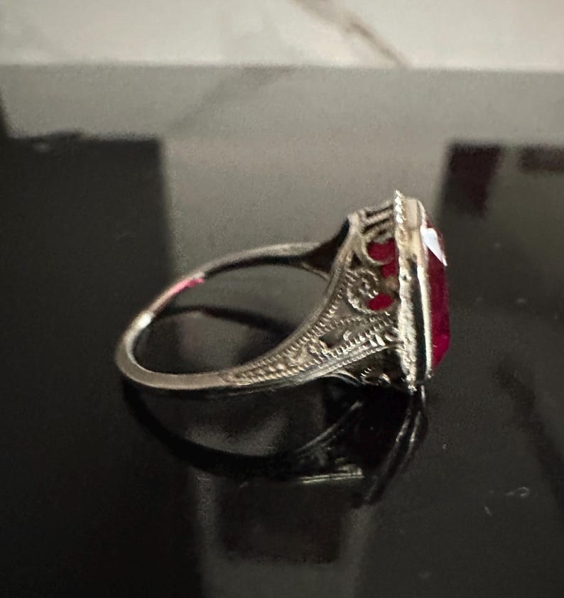Op de afbeelding: Een zilveren ring met een sierlijk filigraanontwerp en een grote, rechthoekige, dieprode edelsteen. De ring is geplaatst tegen een donkere achtergrond, waardoor de ingewikkelde details van het metaalwerk en de levendige kleur van de steen worden benadrukt.
