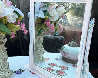 May include: A white vanity mirror with a decorative, ornate frame. The frame is painted with blue and pink accents. The mirror is standing on a table with a floral pattern.