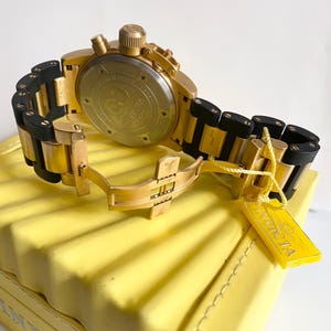 May include: A gold-toned Invicta chronograph watch with black accents on the band, resting on a yellow surface. The watch features a detailed dial and a yellow tag with the Invicta logo. The watch band is a combination of gold and black.