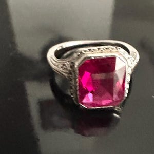 Op de afbeelding: Een zilveren ring met een grote, rechthoekige, felroze edelsteen. De ring heeft een sierlijke, gedetailleerde band met een vintage design. De edelsteen is het middelpunt en reflecteert een levendige roze kleur.