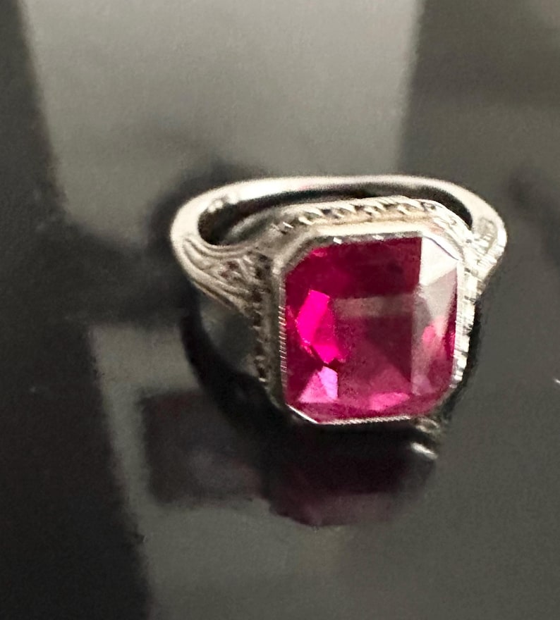 Op de afbeelding: Een zilveren ring met een grote, rechthoekige, felroze edelsteen. De ring heeft een sierlijke, gedetailleerde band met een vintage design. De edelsteen is het middelpunt en reflecteert een levendige roze kleur.