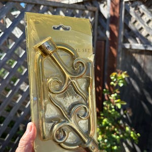 May include: A gold-coloured metal curtain rod holder set in its original packaging. The holder features an ornate, scrolled design. The packaging is a gold-coloured cardboard with the words "HOME" and "WINDOW" visible.