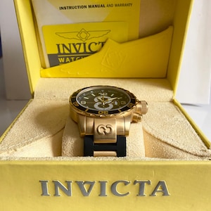 May include: A gold-toned Invicta watch with a black dial and a black rubber strap is displayed inside a yellow box. The watch features a gold bezel and a detailed face with luminous hands and markers. The box has the Invicta logo.