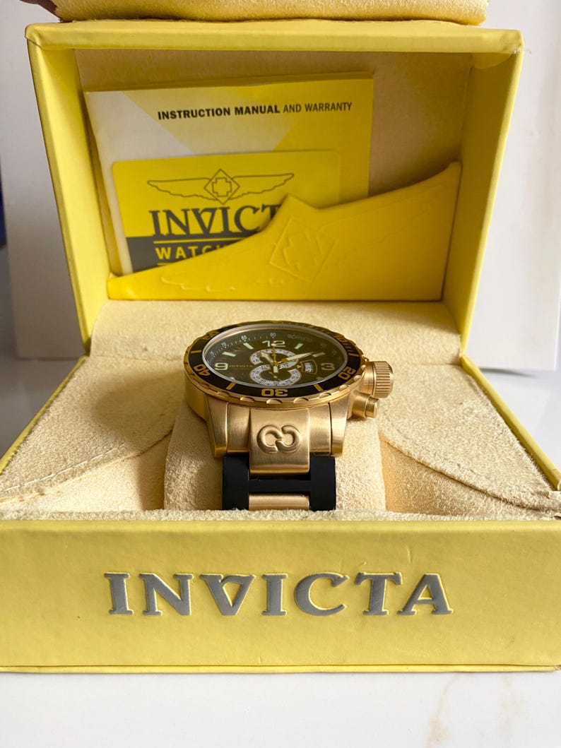 May include: A gold-toned Invicta watch with a black dial and a black rubber strap is displayed inside a yellow box. The watch features a gold bezel and a detailed face with luminous hands and markers. The box has the Invicta logo.
