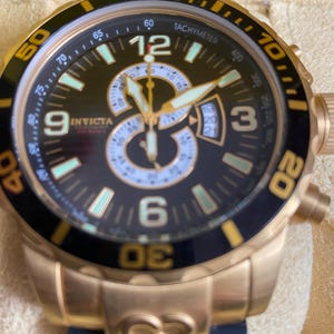May include: A gold-toned Invicta watch with a black dial and a tachymeter bezel. The watch features luminous hands and markers, a date display, and a black and blue band. The watch has a large face with the number 12 prominently displayed.