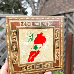 Puede incluir: Caja cuadrada de madera con un borde con incrustaciones decorativas. El centro presenta un mapa de Líbano en rojo, con un cedro verde y la palabra "LEBANON" en blanco. El fondo es un material nacarado.