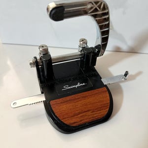 Könnte beinhalten: Ein schwarzer und silberner Swingline-Locher mit Holzmaserung. Der Locher hat einen silbernen Griff und eine Papierführung. Der Text "Swingline" ist auf der Vorderseite aufgedruckt.