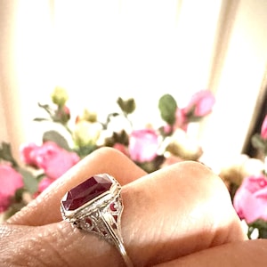 Op de afbeelding: Een zilveren ring met een grote, rechthoekige, dieprode edelsteen. De ring heeft gedetailleerd filigraanwerk op de band en kleinere rode stenen. De achtergrond is vervaagd met roze bloemen.