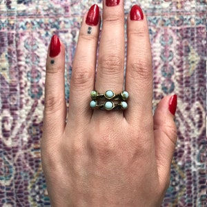 Puede incluir: Un anillo de oro con cinco piedras turquesa dispuestas en fila. El anillo se lleva en el dedo medio de una mano con esmalte de uñas rojo.