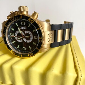 May include: Gold-tone Invicta watch with a black dial and a black and gold band. The watch features a tachymeter and a date display. The watch is resting on a yellow surface.