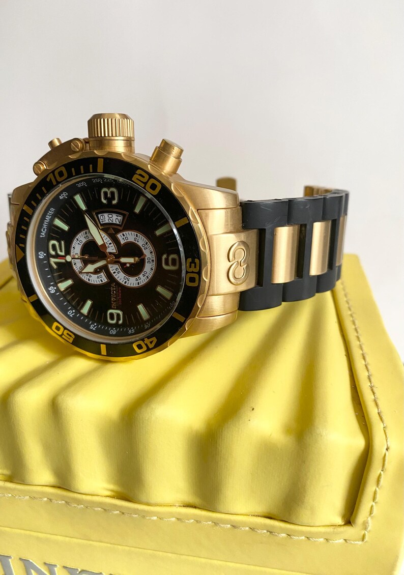 May include: Gold-tone Invicta watch with a black dial and a black and gold band. The watch features a tachymeter and a date display. The watch is resting on a yellow surface.
