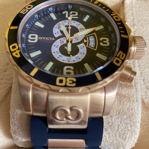 May include: A gold-toned Invicta chronograph watch with a black dial, featuring luminous hands and markers. The watch has a rotating bezel with yellow accents and a blue rubber strap. The watch face displays the Invicta logo and various sub-dials.