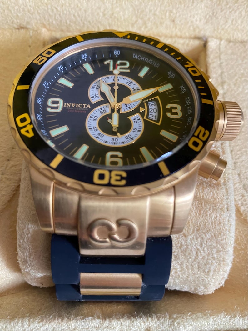 May include: A gold-toned Invicta chronograph watch with a black dial, featuring luminous hands and markers. The watch has a rotating bezel with yellow accents and a blue rubber strap. The watch face displays the Invicta logo and various sub-dials.