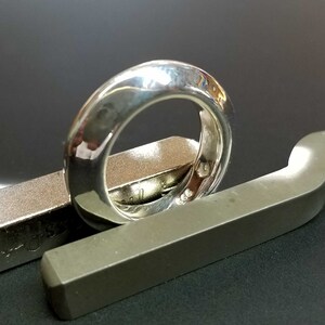 Könnte beinhalten: Ein polierter Silberring mit einem abgerundeten Band. Der Ring liegt auf einer grauen Metalloberfläche. Der Ring hat eine glatte, reflektierende Oberfläche, und das Metall scheint mit Markierungen versehen zu sein.