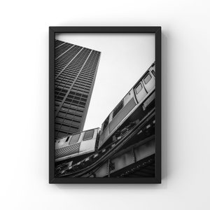 May include: A black and white photograph of a train traveling through a city. The train is seen from below, with the tracks and the underside of the train visible. A tall building is visible in the background.