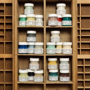 May include: A wooden shelf unit with multiple rows of small glass jars filled with different colored paint. Each jar has a white lid and a label that reads "Fusion".
