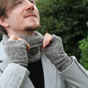 May include: A gray tweed knit cowl and fingerless gloves set. The cowl is around the neck, and the gloves cover the hands. The set is made of a speckled gray yarn. The person in the image is wearing a gray coat.