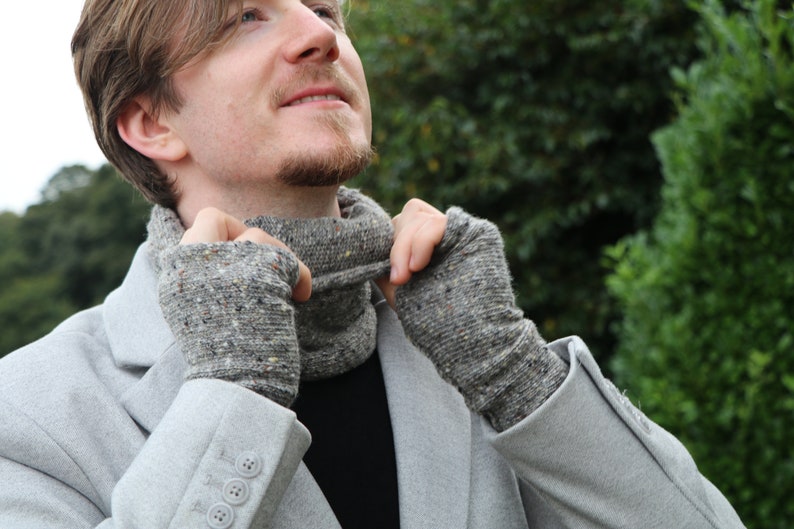 May include: A gray tweed knit cowl and fingerless gloves set. The cowl is around the neck, and the gloves cover the hands. The set is made of a speckled gray yarn. The person in the image is wearing a gray coat.