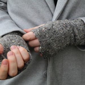 May include: Close-up of gray knitted fingerless gloves with flecks of color. The gloves are worn with a gray coat. The gloves are made of a soft, textured yarn, perfect for keeping hands warm.