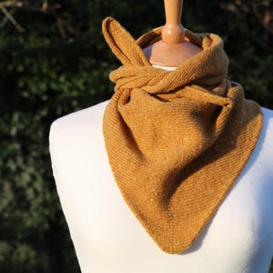 Puede incluir: Una bufanda triangular de color amarillo mostaza con una textura de punto fino, drapeada alrededor del cuello de un maniquí blanco. La bufanda presenta un diseño de nudo simple en la parte delantera, mostrando una construcción de hilo uniforme.