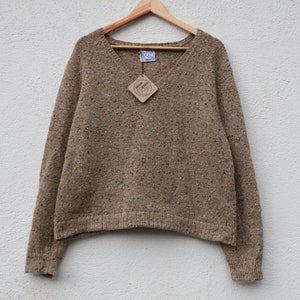 May include: A brown speckled v-neck sweater with long sleeves. The sweater is hanging on a wooden hanger against a white wall. The sweater has a small brown tag with the text "Irish Tweed" on it.