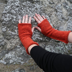 May include: A pair of orange fingerless gloves made of a textured knit fabric. The gloves are a vibrant shade of orange with flecks of other colors. The gloves are shown against a textured stone background.