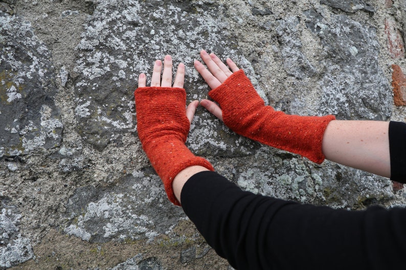 May include: A pair of orange fingerless gloves made of a textured knit fabric. The gloves are a vibrant shade of orange with flecks of other colors. The gloves are shown against a textured stone background.