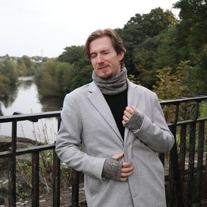 May include: A light gray blazer, black shirt, and gray knit accessories are worn by a person standing on a bridge. The person is wearing a gray knit cowl and matching fingerless gloves. The background features a river and trees.