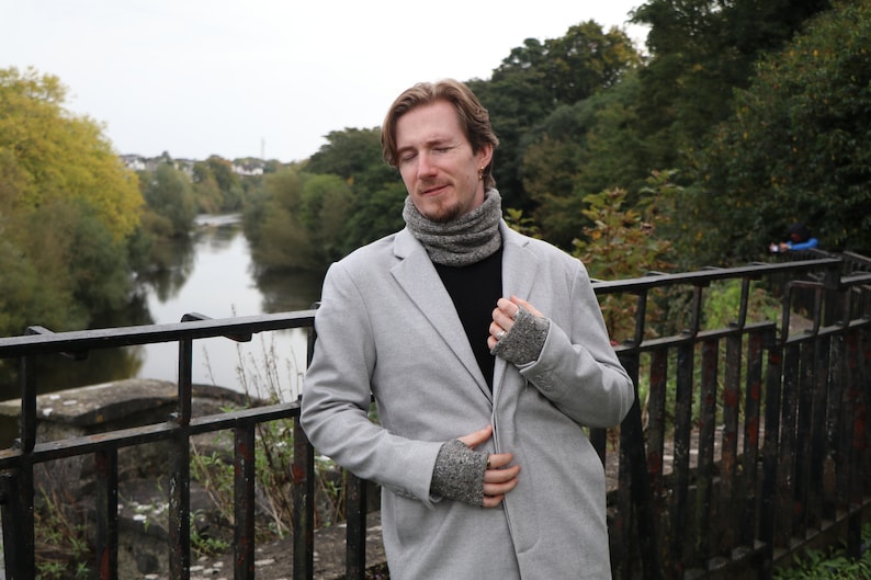 May include: A light gray blazer, black shirt, and gray knit accessories are worn by a person standing on a bridge. The person is wearing a gray knit cowl and matching fingerless gloves. The background features a river and trees.